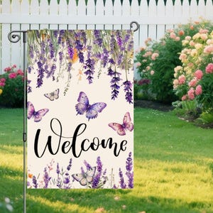 May include: A garden flag with a cream background features purple lavender and butterflies. The word "Welcome" is written in a cursive font. The flag is displayed on a black metal stand in a garden setting.