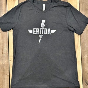May include: Dark gray t-shirt with a white graphic. The graphic features the word "EBITDA" with a lightning bolt and wings design. The shirt is made of a soft material.