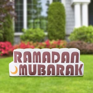 May include: A white and brown yard sign with the words "RAMADAN MUBARAK" in a bold, stylized font. The sign includes a crescent moon graphic. The sign is placed on a green lawn in front of a house.