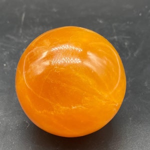 以下が含まれることがあります： 鮮やかなオレンジ色の滑らかで磨かれた球体。表面には繊細で半透明な内部の筋模様と、色の濃淡の自然な変化が見られ、丸みを帯びた形状全体に光沢のある反射する外観を作り出しています。