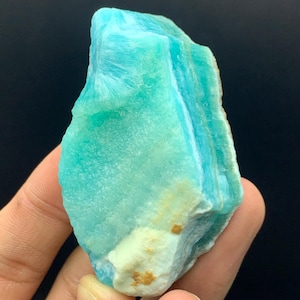 Può includere: Una gemma di amazzonite grezza, che mostra una miscela di tonalità turchesi e blu con venature bianche. La pietra ha un aspetto naturale, non lucido, con una base color beige chiaro.