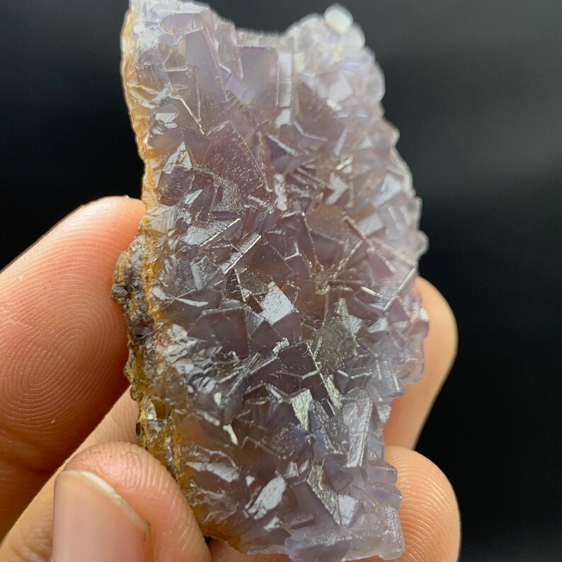 K&ouml;nnte beinhalten: Ein roher, nat&uuml;rlicher Fluorit-Kristallcluster. Der Kristall zeigt ein komplexes geometrisches Muster mit Lila- und Lavendelt&ouml;nen. Die Kanten haben einen br&auml;unlich-gelben Farbton. Der Kristall wird in einer Hand vor dunklem Hintergrund gehalten.