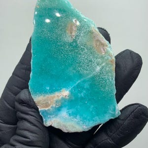 May include: A vibrant, teal-colored mineral specimen with a rough, textured surface. The stone has areas of light brown and white, and is held in a black gloved hand. The overall shape is irregular, with a natural, organic aesthetic.