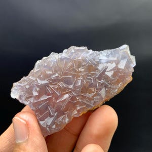 K&ouml;nnte beinhalten: Eine Hand h&auml;lt einen rohen, ungeschliffenen Fluoritkristall. Der Kristall weist ein komplexes geometrisches Muster mit Schattierungen von Lila, Wei&szlig; und Braun auf. Die rauen Kanten und die nat&uuml;rliche Formation sind sichtbar.