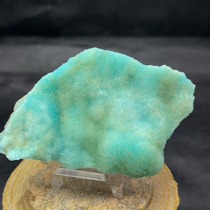 May include: A vibrant, turquoise mineral specimen with a textured surface. The stone is displayed on a clear stand, showcasing its natural form and color variations. The background is dark, highlighting the mineral's bright hue.