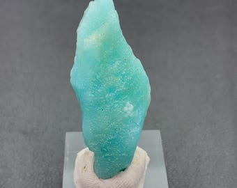 Puede incluir: Un vibrante espécimen mineral de color turquesa con una superficie rugosa y texturizada. Montado sobre una base rectangular transparente con un soporte blanco texturizado. La forma general es irregular y orgánica, con variaciones de color.
