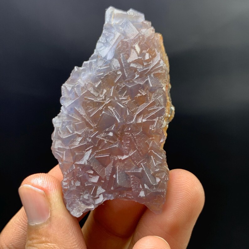 K&ouml;nnte beinhalten: Ein roher, nat&uuml;rlicher Fluoritkristall mit einem komplexen, geometrischen Muster. Der Kristall zeigt eine Mischung aus transluzenten violetten und braunen Farbt&ouml;nen mit einer rauen, strukturierten Oberfl&auml;che. Das Mineral wird in einer Hand vor dunklem Hintergrund gehalten.