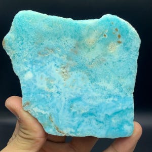 May include: A vibrant, irregularly shaped piece of larimar gemstone. The stone displays a captivating blend of light and medium blue hues, with subtle white and brown veining throughout. The surface has a natural, textured appearance, showcasing the stone's unique character.