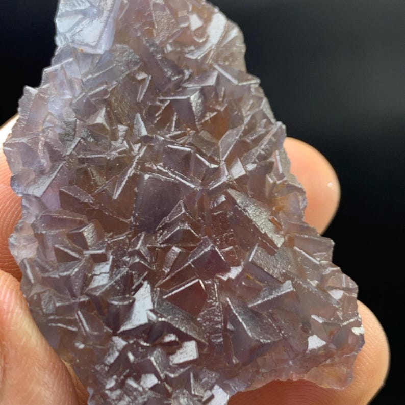 K&ouml;nnte beinhalten: Nahaufnahme eines rohen, nat&uuml;rlichen Fluoritkristalls. Der Kristall hat eine komplexe, geometrische Struktur mit vielen kleinen, quadratischen und dreieckigen Facetten. Die Farbe ist eine Mischung aus Lila und Braun, mit einer durchscheinenden Qualit&auml;t.