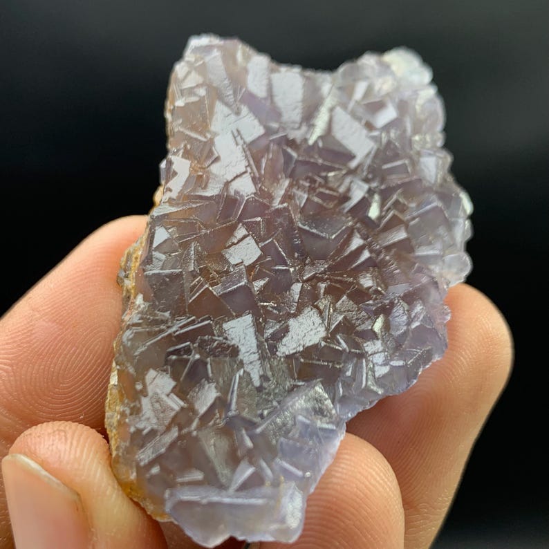 K&ouml;nnte beinhalten: Nahaufnahme einer Fluorit-Mineralprobe. Der Fluorit hat eine kristalline Struktur mit Lila- und Wei&szlig;t&ouml;nen. Die Kanten des Minerals sind hellbraun. Der Hintergrund ist schwarz.