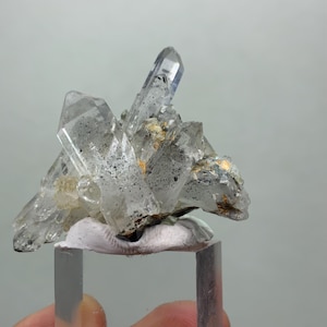 May include: A cluster of clear quartz crystals with dark inclusions and a few gold-colored accents. The crystals are mounted on a white base and a clear rectangular stand. The quartz formation has a natural, rough texture and is set against a neutral background.