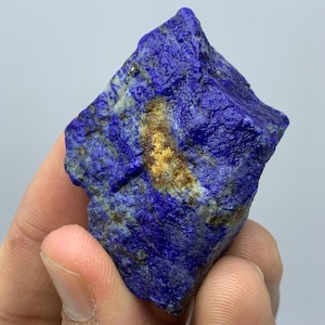 Op de afbeelding: Een diepblauwe lapis lazuli steen met gouden insluitsels. De natuurlijke edelsteen heeft een getextureerd oppervlak en een onregelmatige vorm, die zijn ruwe, ongepolijste staat laat zien. De steen wordt in een hand gehouden.