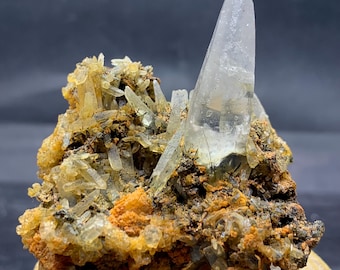 Può includere: Un gruppo di cristalli di quarzo trasparente con un grande cristallo appuntito che emerge da una base di cristalli più piccoli, giallastri e roccia marrone. Il quarzo è traslucido e la base ha sfumature arancioni e marroni.