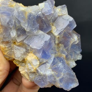 May include: A close-up of a cluster of blue and clear fluorite crystals. The crystals have a cubic structure and are partially translucent, with some areas appearing more opaque. The base of the cluster has a tan color.