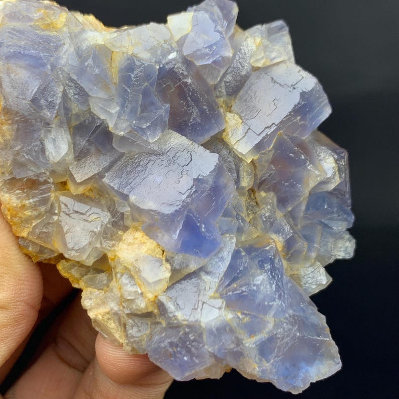 May include: A close-up of a cluster of blue and clear fluorite crystals. The crystals have a cubic structure and are partially translucent, with some areas appearing more opaque. The base of the cluster has a tan color.