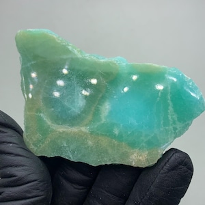 May include: A polished, raw gemstone in shades of teal and green. The stone has a rough, natural edge and a smooth, polished surface. The color transitions from a light green to a deep teal, with areas of lighter color and a hint of brown.