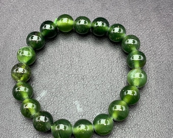 May include: A green beaded bracelet. The bracelet is made of round, polished beads in varying shades of green. The beads have a reflective, glossy surface. The bracelet is displayed on a dark gray surface.