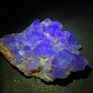 May include: A cluster of vibrant, crystalline formations in shades of deep blue and purple. The mineral specimen has a rough, textured surface with sharp, geometric facets. The base is a light tan color, providing a contrast to the intense blue crystals.