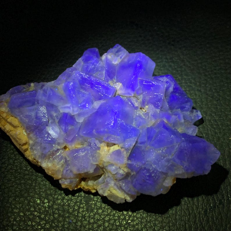 May include: A cluster of vibrant, crystalline formations in shades of deep blue and purple. The mineral specimen has a rough, textured surface with sharp, geometric facets. The base is a light tan color, providing a contrast to the intense blue crystals.