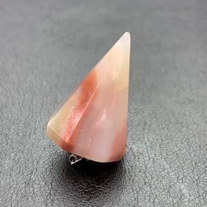 May include: A triangular brooch made from polished stone, displaying bands of red, pink, and cream hues. The brooch has a silver pin for fastening. The stone has a smooth, glossy finish. The brooch is approximately 5 cm in length.