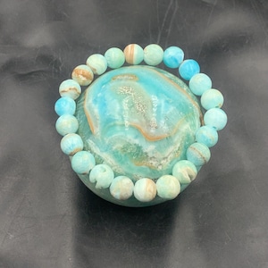 May include: A beaded bracelet featuring round, light blue and tan beads. The bracelet is resting on a round, turquoise stone with a marbled pattern. The beads are strung together, forming a complete circle.