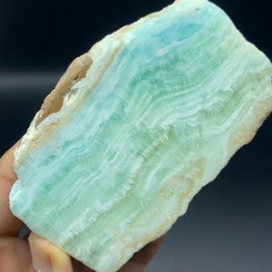 May include: A polished slab of Caribbean calcite featuring wavy, banded layers of light blue, mint green, and creamy white. The mineral specimen displays a translucent, crystalline texture with natural, rough, tan-coloured edges along one side.