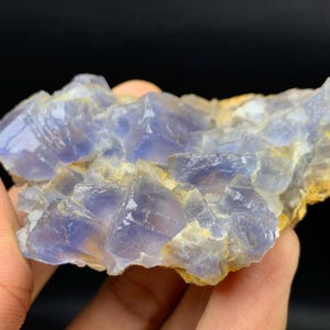 May include: A close-up of a blue and white fluorite mineral specimen. The fluorite crystals have a translucent quality, with a mix of light and dark blue hues. The mineral sits on a light brown matrix, with a dark background.