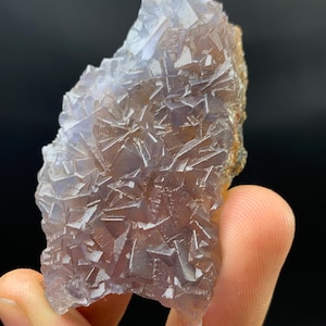 K&ouml;nnte beinhalten: Nahaufnahme einer Fluorite-Mineralprobe. Die Kristallstruktur zeigt ein komplexes Muster geometrischer Formen in Lila-, Braun- und Klart&ouml;nen. Das Mineral wird von den Fingern einer Person vor einem schwarzen Hintergrund gehalten.