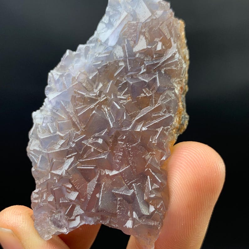 K&ouml;nnte beinhalten: Nahaufnahme einer Fluorite-Mineralprobe. Die Kristallstruktur zeigt ein komplexes Muster geometrischer Formen in Lila-, Braun- und Klart&ouml;nen. Das Mineral wird von den Fingern einer Person vor einem schwarzen Hintergrund gehalten.