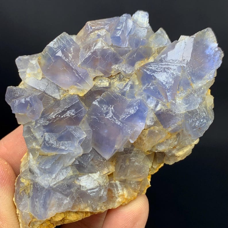 May include: A natural fluorite mineral specimen featuring clusters of translucent, blue-purple cubic crystals. The crystals have a glassy luster and are set against a matrix of tan and brown rock. The specimen is a unique example of geological formations.