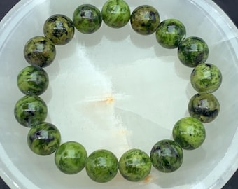 May include: A beaded bracelet featuring round, green gemstones with dark spots. The bracelet is displayed on a white, bowl-shaped surface. The gemstones have a polished appearance and are strung together to form a circular shape.