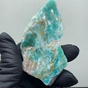 May include: A rough-cut, natural stone specimen with a blend of teal, green, and tan hues. The stone has a crystalline structure and a rugged, uneven surface. The colors are swirled and layered, creating a unique pattern. The stone is held by a gloved hand.