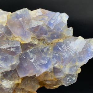 May include: Close-up of a fluorite mineral specimen. The crystalline structure displays a mix of pale purple and clear, translucent sections. The edges of the crystals have a golden-brown hue, with a dark background.