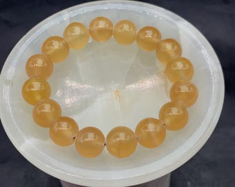 Può includere: Un braccialetto di perline giallo dorato. Il braccialetto è composto da perline rotonde e lucide, infilate per formare un cerchio completo. Le perline hanno una calda tonalità ambrata e sono esposte su una superficie bianca e circolare.