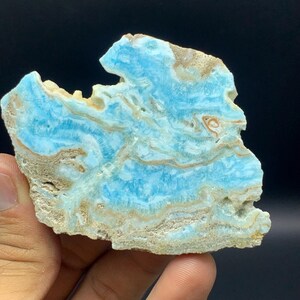 May include: A polished slab of blue larimar stone featuring intricate swirling patterns of sky blue, white, and tan mineral inclusions. The specimen has a rough, unpolished edge along the top and bottom, showcasing natural textures and earthy tones.