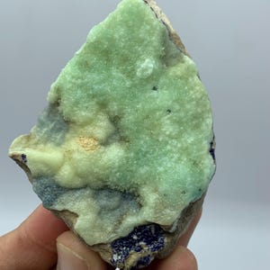 May include: A rough-cut mineral specimen with a light green, textured surface. The stone has areas of pale yellow and dark blue, with a rough, uneven shape. The mineral is held in a hand, showing its natural form and color variations.