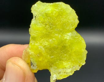 May include: A vibrant, irregularly shaped, yellow-green mineral specimen. The rough, textured surface of the stone is held against a dark background. The mineral's color is a bright, eye-catching hue.
