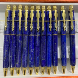 May include: A set of eleven ballpoint pens featuring deep blue lapis lazuli stone barrels with gold-tone metal accents. Each pen includes a gold clip with a small embedded clear stone at the top and a textured gold tip for a consistent appearance.