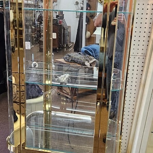 May include: A gold-toned glass display cabinet with multiple shelves. The cabinet has a mirrored back and is made of glass and metal.