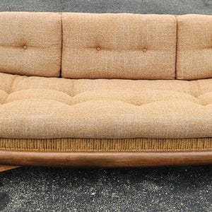 May include: A mid-century modern sofa with a curved, low profile. The sofa features a light brown fabric with button tufting on the seat and back cushions. The wooden frame has a curved design and angled legs.