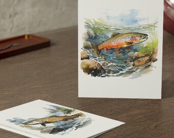May include: Two greeting cards featuring watercolor paintings of fish in a stream. The fish have orange and brown markings. The cards are on a dark wooden surface, with a mirror and a decorative vase in the background.