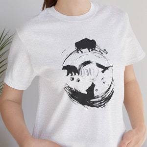 May include: A white t-shirt with a black and white graphic design of a wolf, bear, buffalo, and birds in a circle. The design is stylized and has a brushstroke effect.