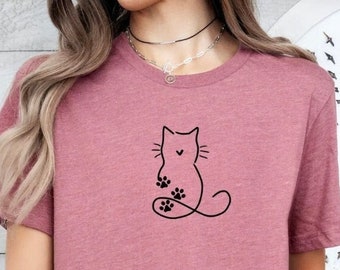 Può includere: Una maglietta a maniche corte color malva con un contorno nero di un gatto con le impronte delle zampe sulla coda.