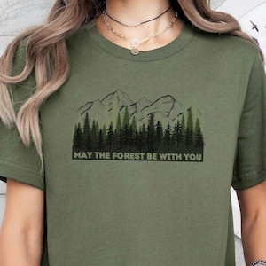 May include: A green t-shirt with a graphic of a mountain range and trees. The text "May the forest be with you" is printed below the graphic.