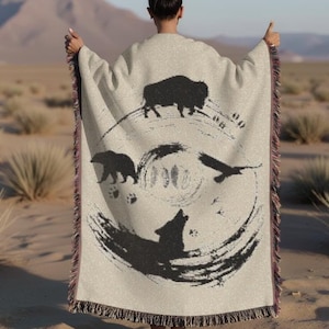 May include: A person wearing a beige woven throw blanket with a black and white design featuring a wolf, a bear, a bird, and a buffalo. The design is circular and includes feathers and paw prints.