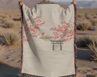 May include: A woven throw blanket with a Japanese-inspired design featuring a mountain, a red sun, a torii gate, and pink maple leaves on a cream background.