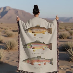 May include: A woven throw blanket featuring three different types of trout: brown, rainbow, and brook. Each fish is depicted in detail with realistic colours and patterns. The blanket is beige with a fringe border.