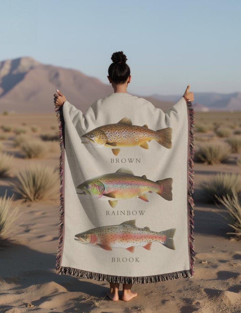 May include: A woven throw blanket featuring three different types of trout: brown, rainbow, and brook. Each fish is depicted in detail with realistic colours and patterns. The blanket is beige with a fringe border.