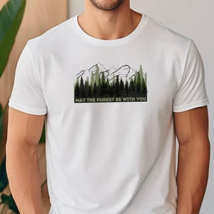 May include: White t-shirt with a green and black mountain and forest graphic. The text "May the forest be with you" is printed below the graphic.