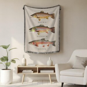 May include: A woven wall tapestry featuring three different types of trout: brown, rainbow, and brook. Each fish is depicted in a realistic style with detailed markings and colours. The tapestry has a fringed border and is hung on a white wall.